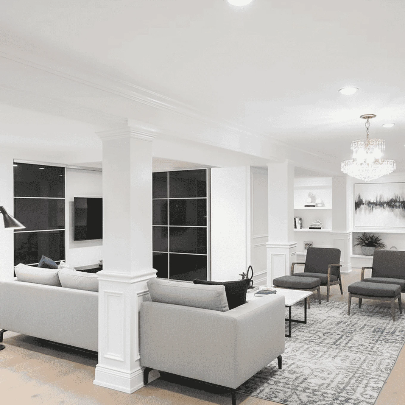 Modern basement renovation in Edgewater, New Jersey featuring a bright lounge area with sectional sofa, accent chairs, chandelier lighting, and built-in shelving.