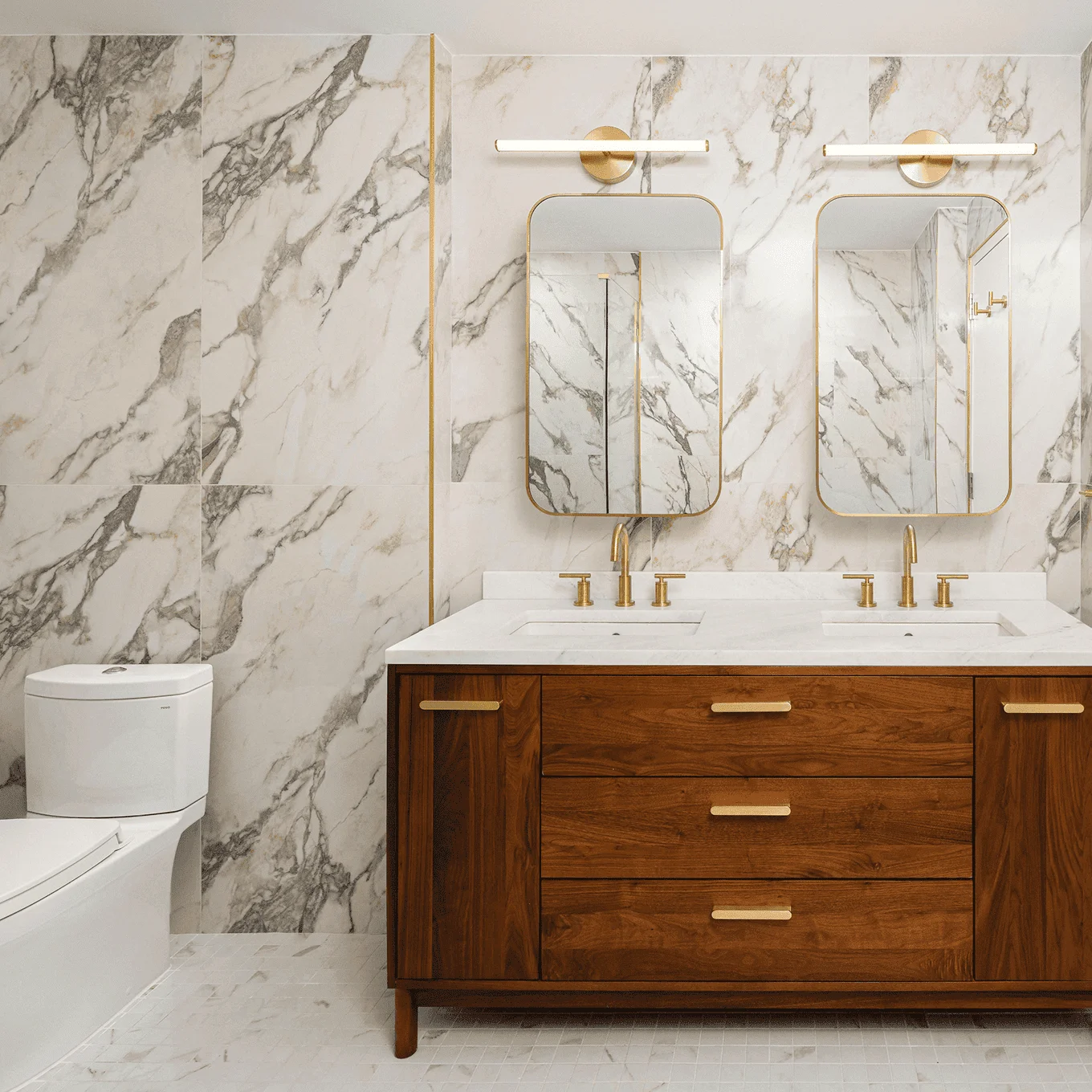 NYC condo bathroom with marble walls, wood double vanity with brass pulls, twin arched mirrors and brass faucets and sconces.