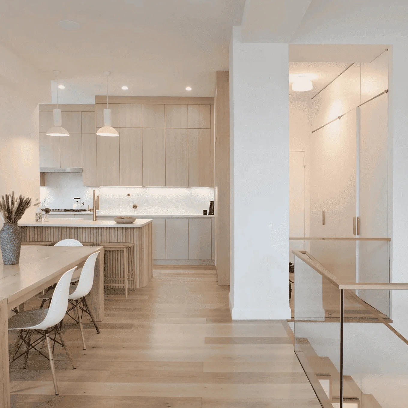 Light-wood kitchen with island and pendants, open dining area and glass-railed staircase in NYC apartment