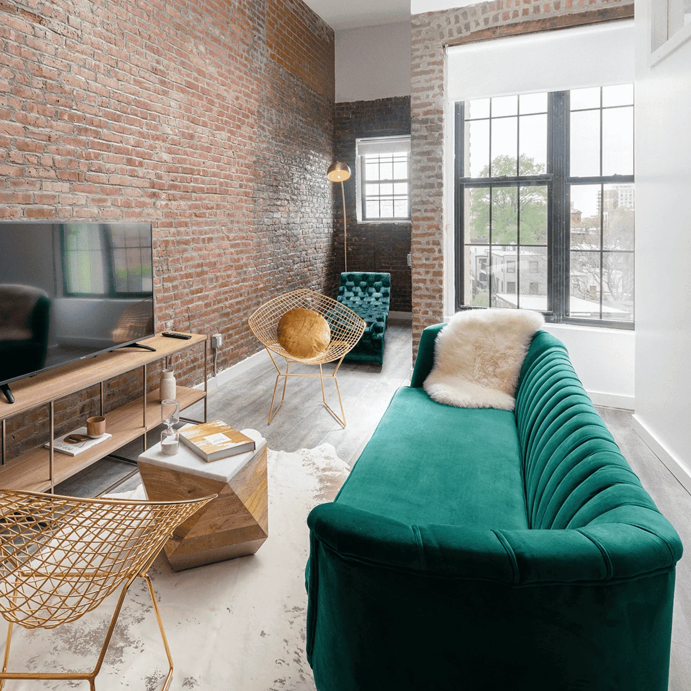 Light-filled living room: brick walls, tall factory windows, green sofa, yellow wire chairs and TV console, Newark Ave apartment.