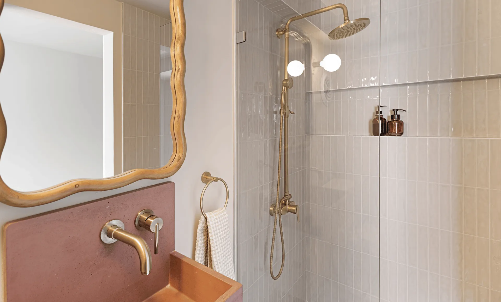 Close-up of brass mirror, wall-mounted brass faucet and matching rain shower against pale vertical tiles.