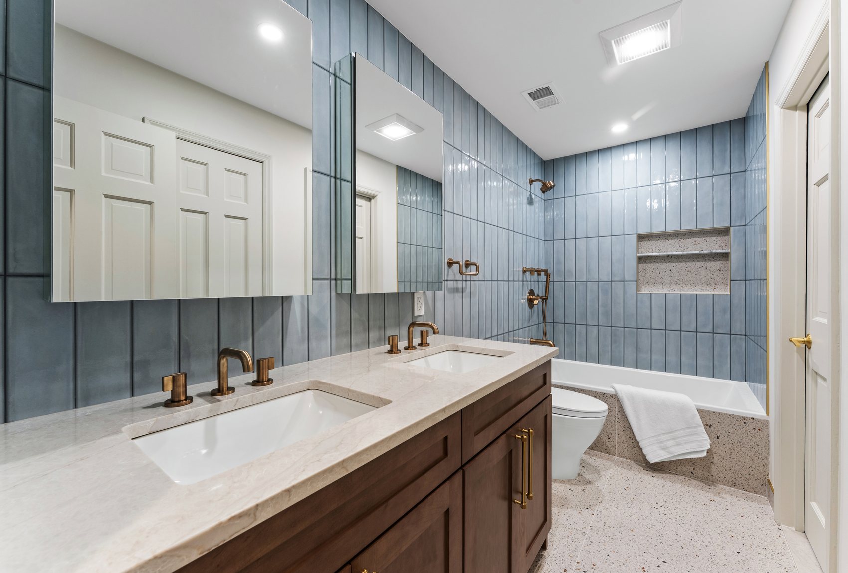 After view of the kids bathroom with blue vertical wall tile, wood vanity, brass fixtures, and a soaking tub.