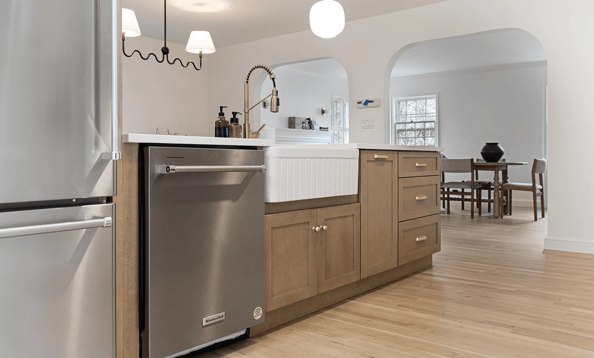Wide view of renovated kitchen featuring farmhouse sink, wood lower cabinets, stainless appliances and arched openings to bright dining area.