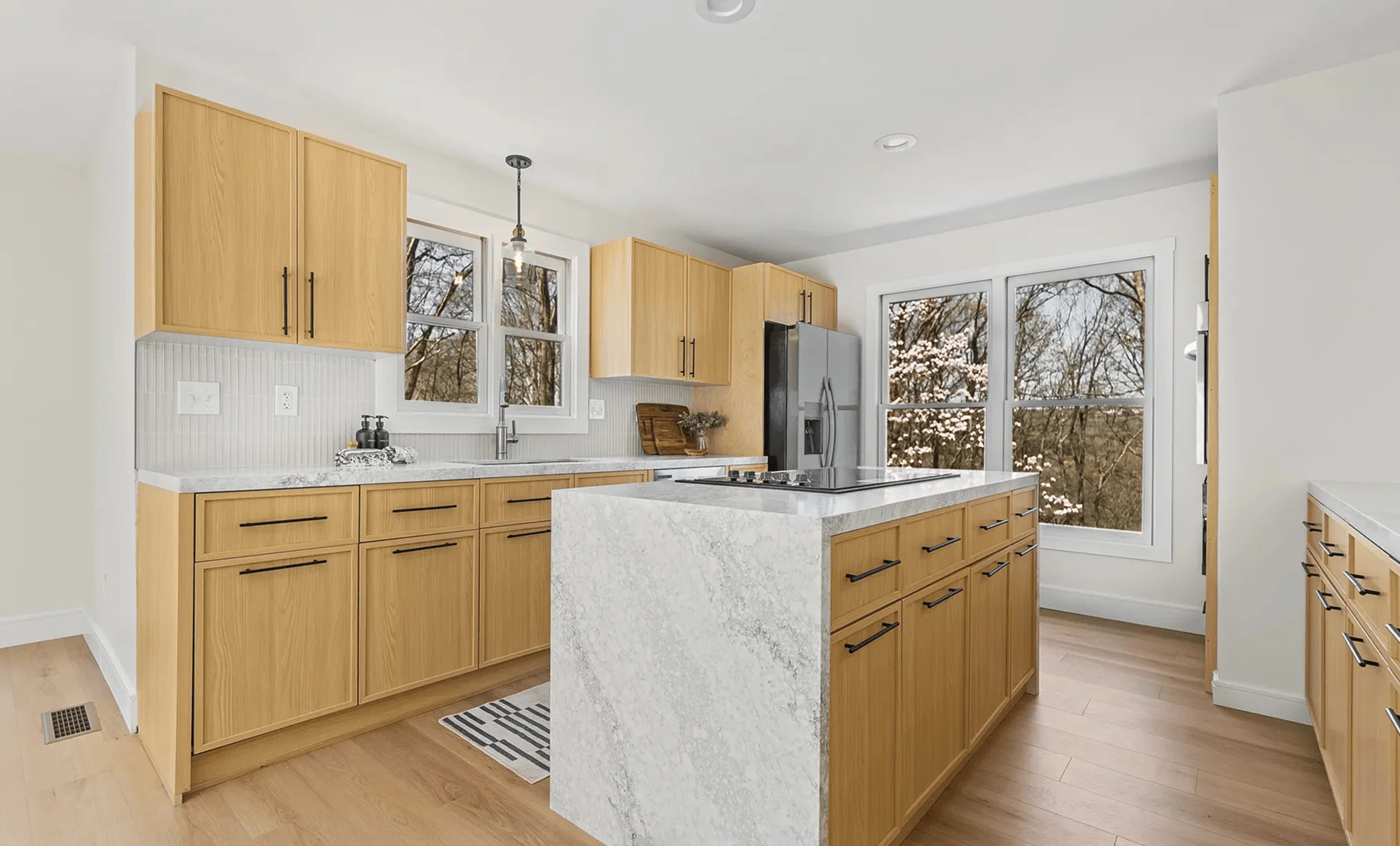 Updated kitchen with quartz waterfall island, vertical tile backsplash, natural wood cabinets and large windows
