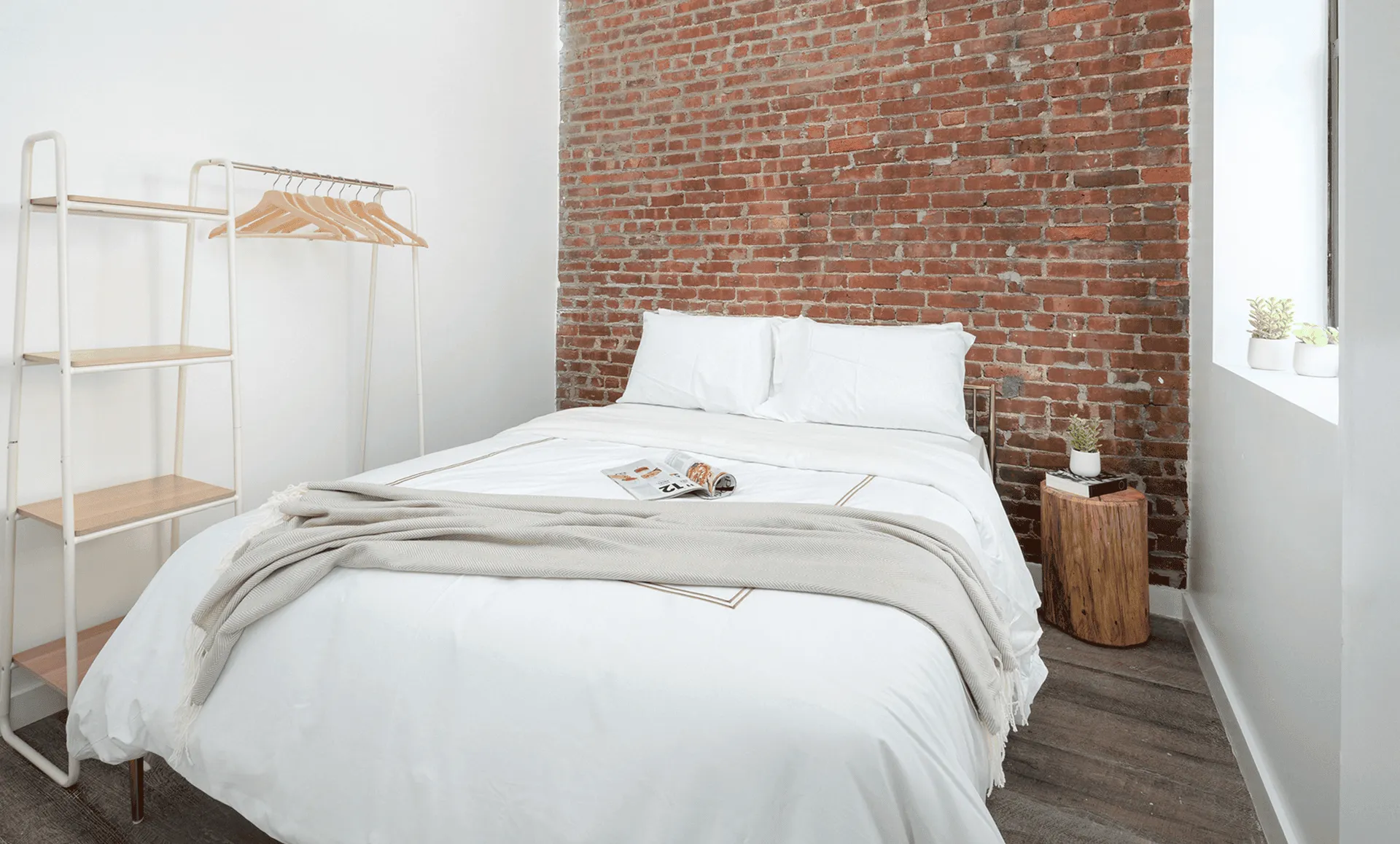 Bedroom with exposed brick accent wall, white bedding, minimalist open wardrobe rack and tree-stump nightstand.