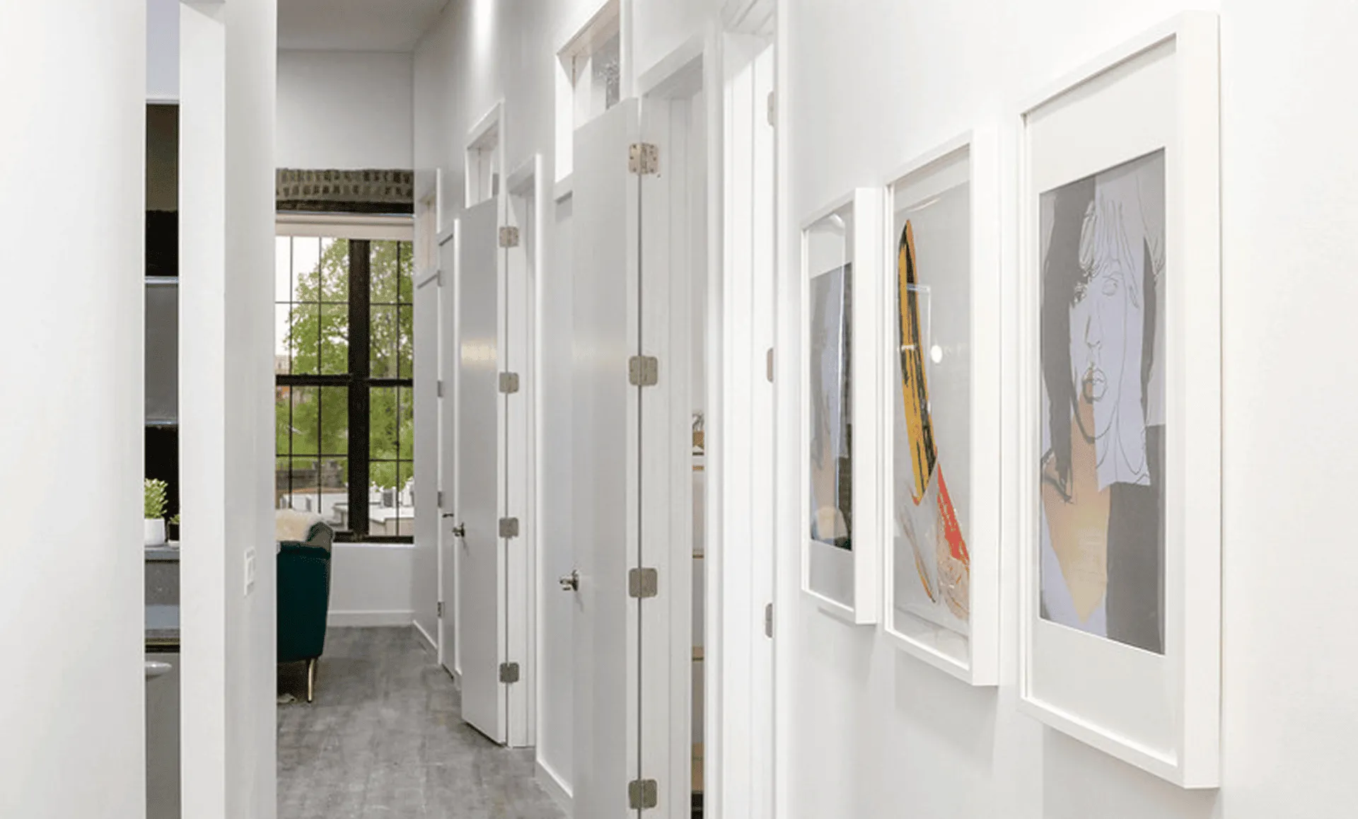Bright hallway lined with bedroom doors and framed art, leading to large industrial window and green sofa beyond.