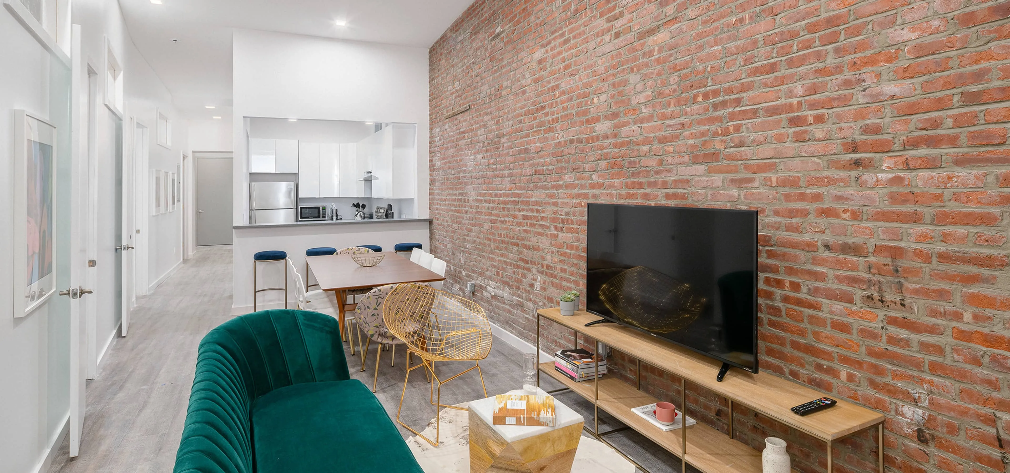 Open-plan Jersey City home with brick feature wall, green velvet sofa, six-seat dining set and pass-through white kitchen.