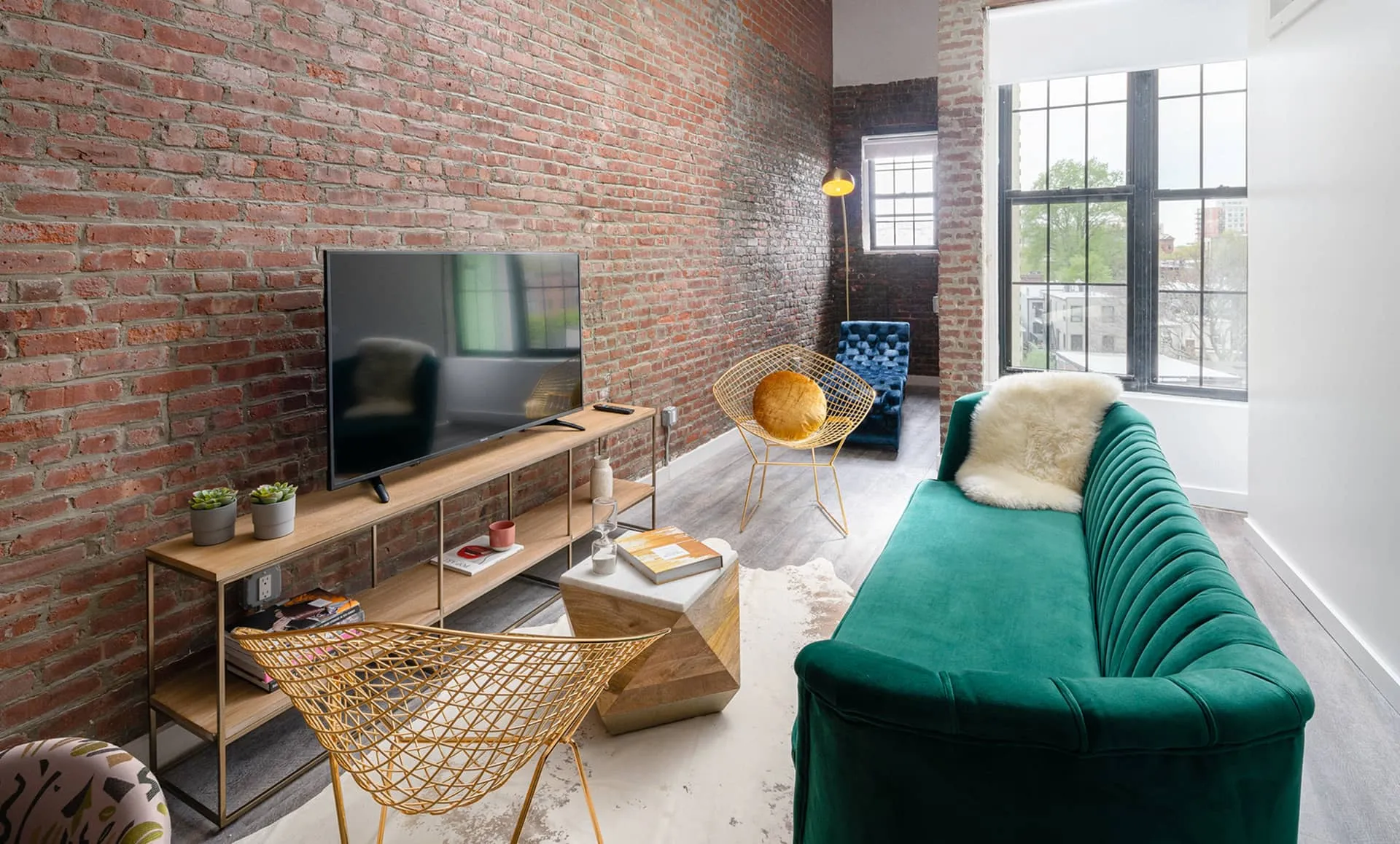 Light-filled living room: brick walls, tall factory windows, green sofa, yellow wire chairs and TV console, Newark Ave apartment.