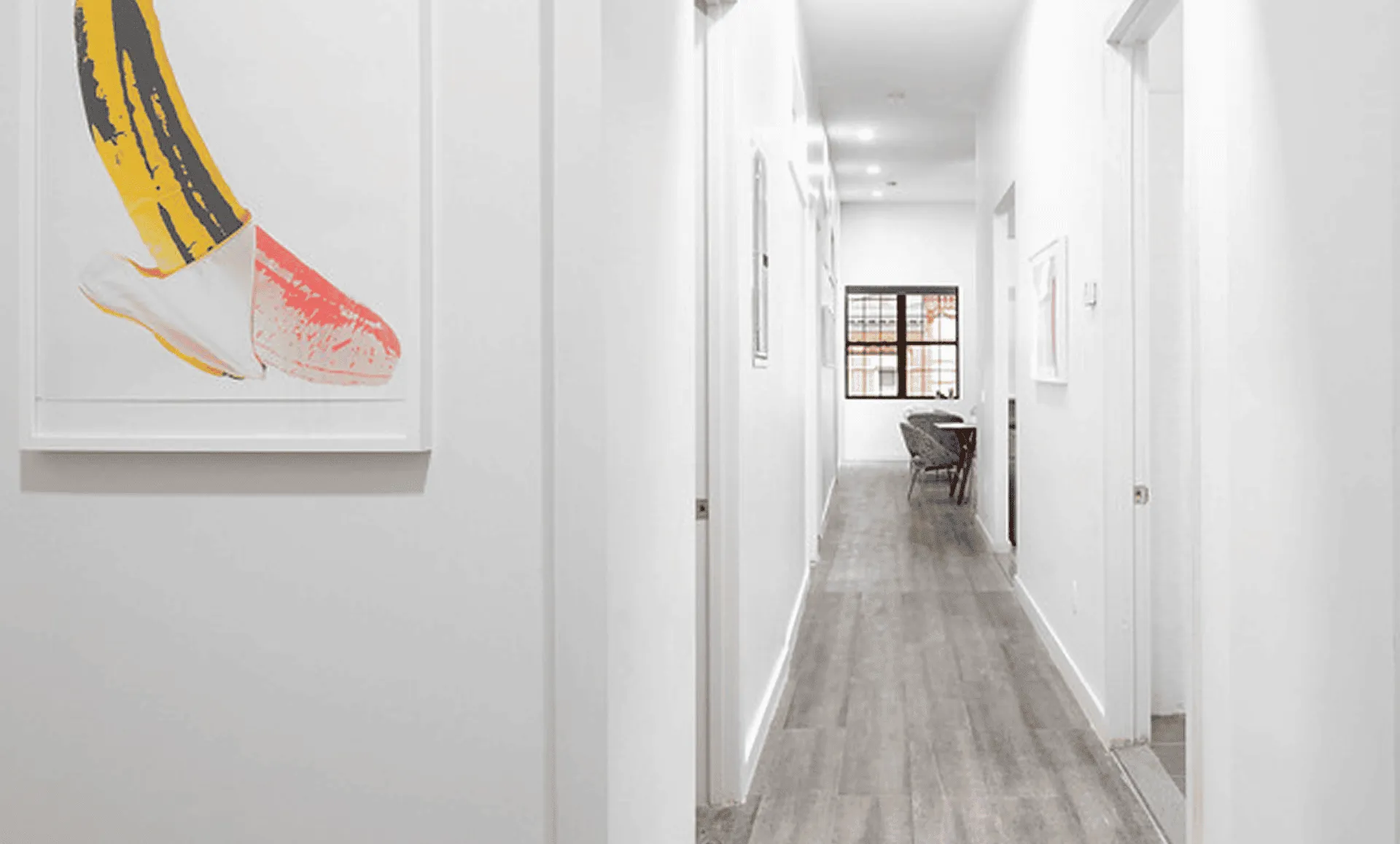 Long white corridor with Warhol banana print, gray plank flooring and view toward dining nook in Newark Ave apartment.