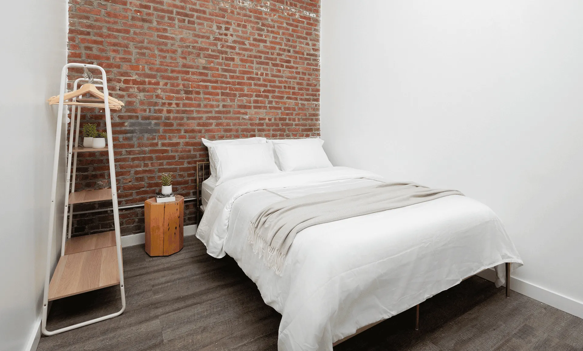 Second minimalist bedroom: brick wall backdrop, white bedding, beige throw and ladder-style shelving rack