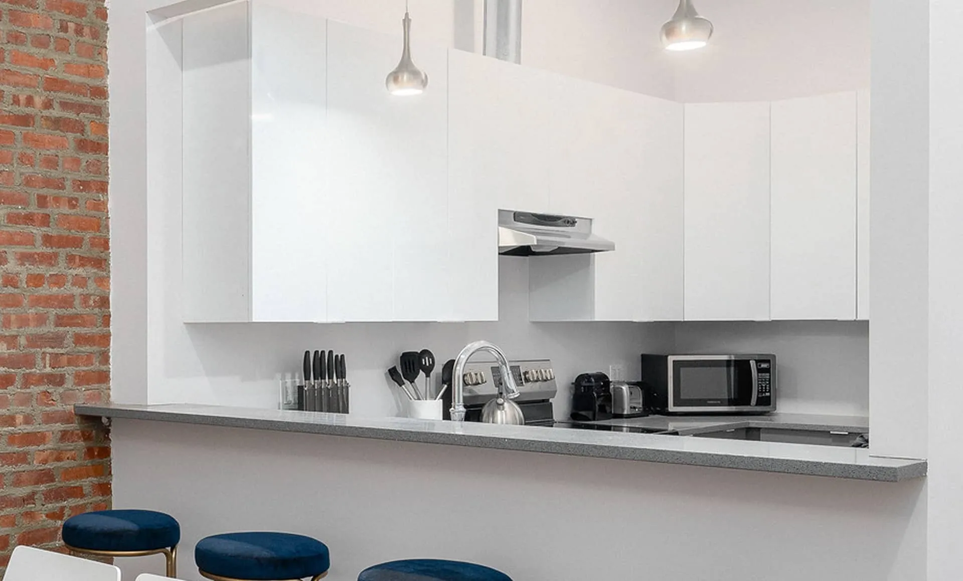 Breakfast bar in Jersey City apartment with gray countertop, white cabinets and blue velvet bar stools against brick wall.