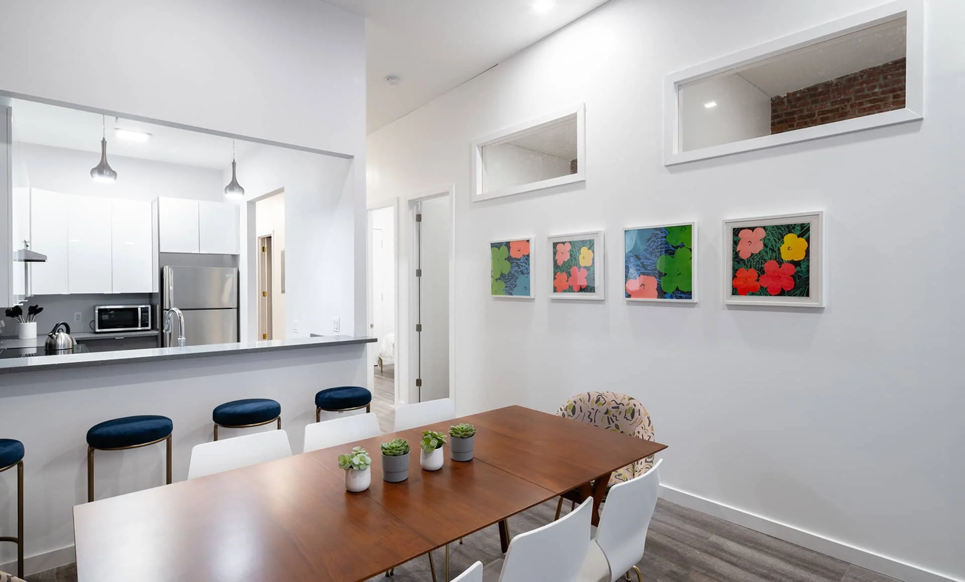 Dining space with walnut table, succulents centerpiece, pop-art floral prints and view into glossy white Jersey City kitchen.