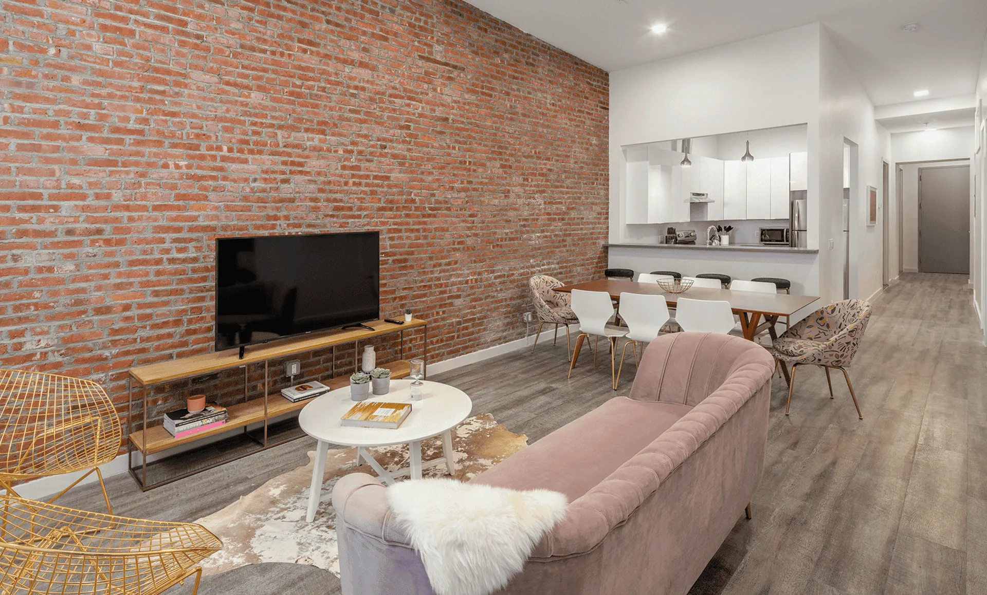 Jersey City apartment on Newark Ave with exposed-brick feature wall, pink sofa, TV console, six-seat dining table and open pass-through kitchen.
