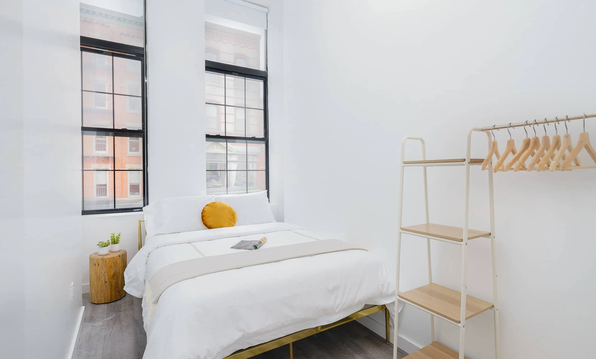 Light-filled bedroom with two tall windows, white walls, gold accent pillow and open shelving wardrobe in Jersey City apartment.
