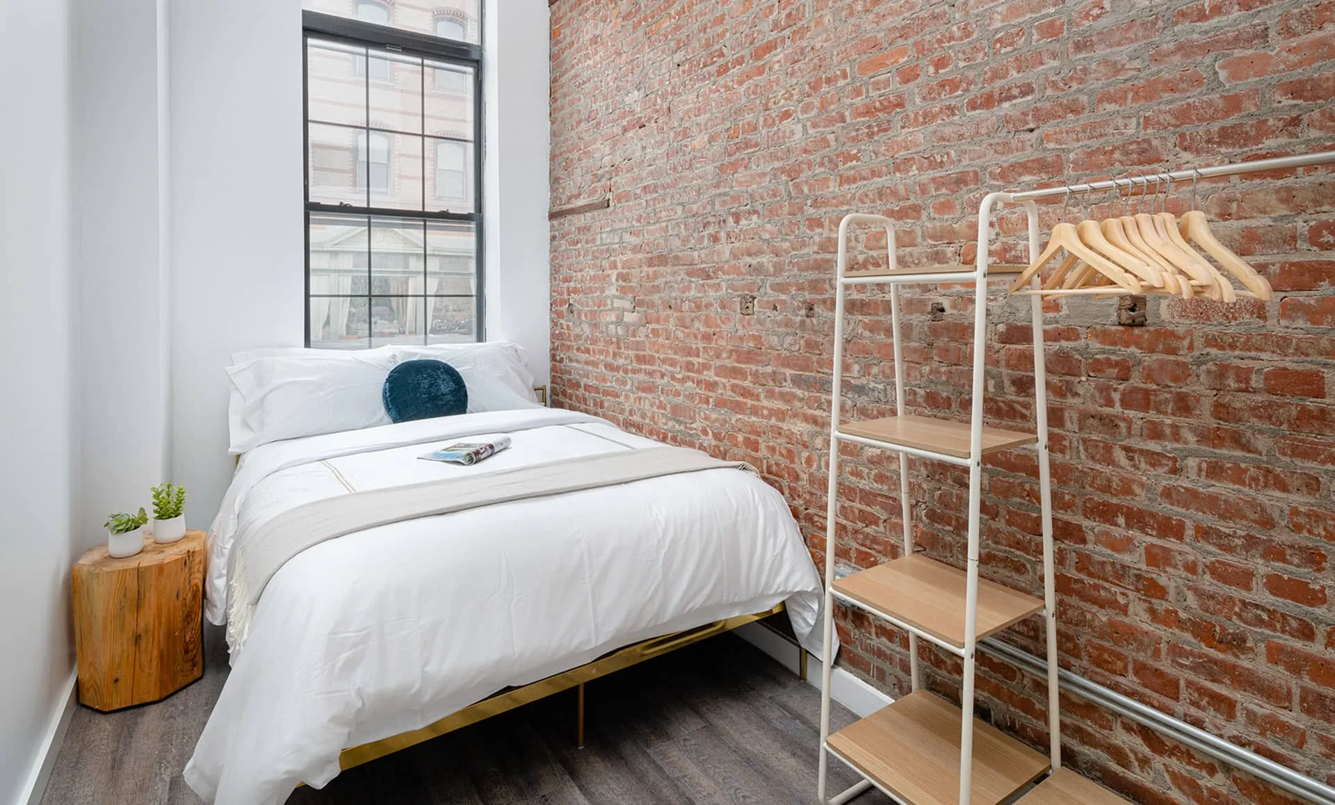 Slim bedroom showing exposed brick wall, large window, white bed with teal pillow and freestanding shelving rack.