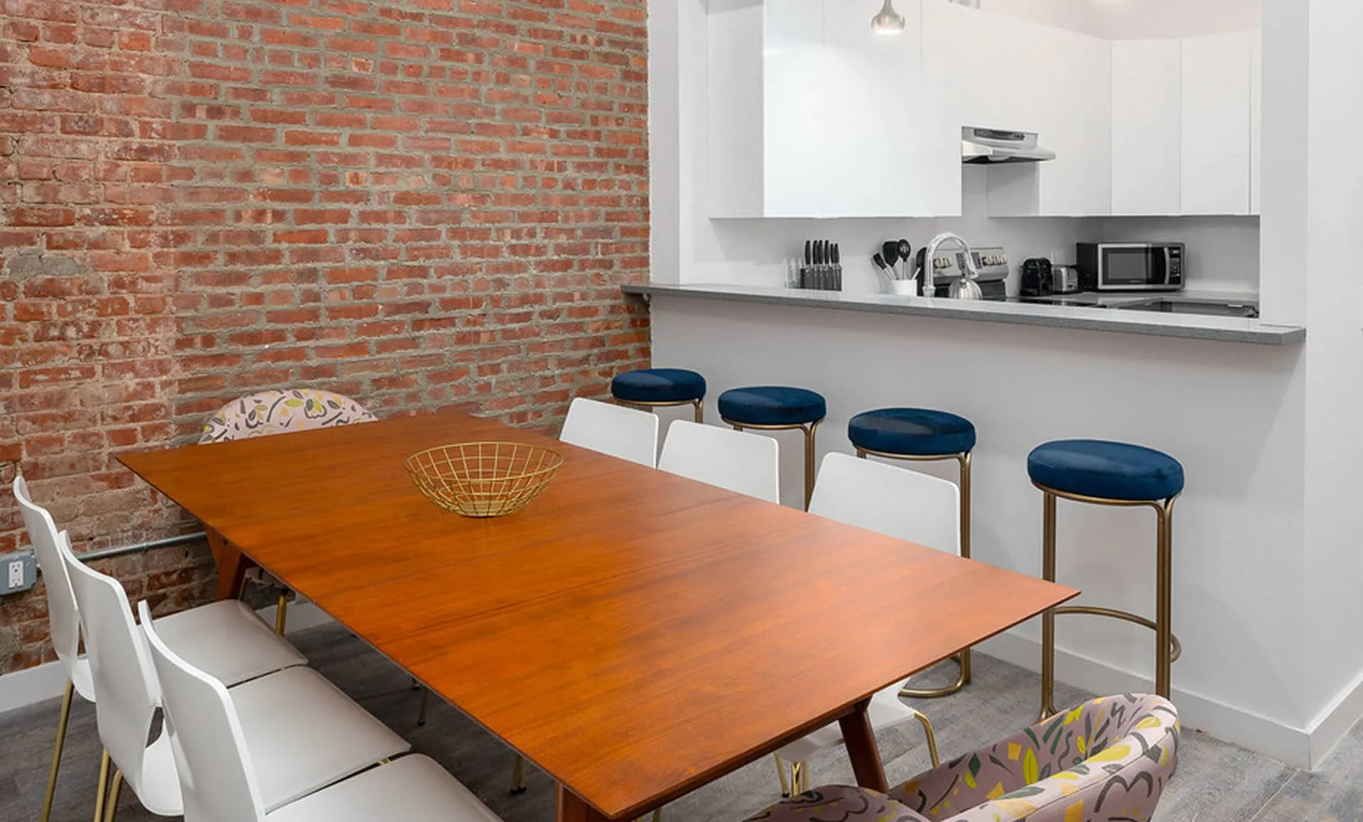 Walnut dining table for eight set beside exposed brick in Newark Ave loft, facing gray breakfast bar with blue stools.