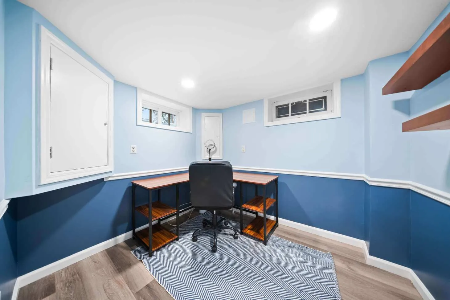 Basement office with two-tone blue walls, wood desk, black chair, and floating shelves