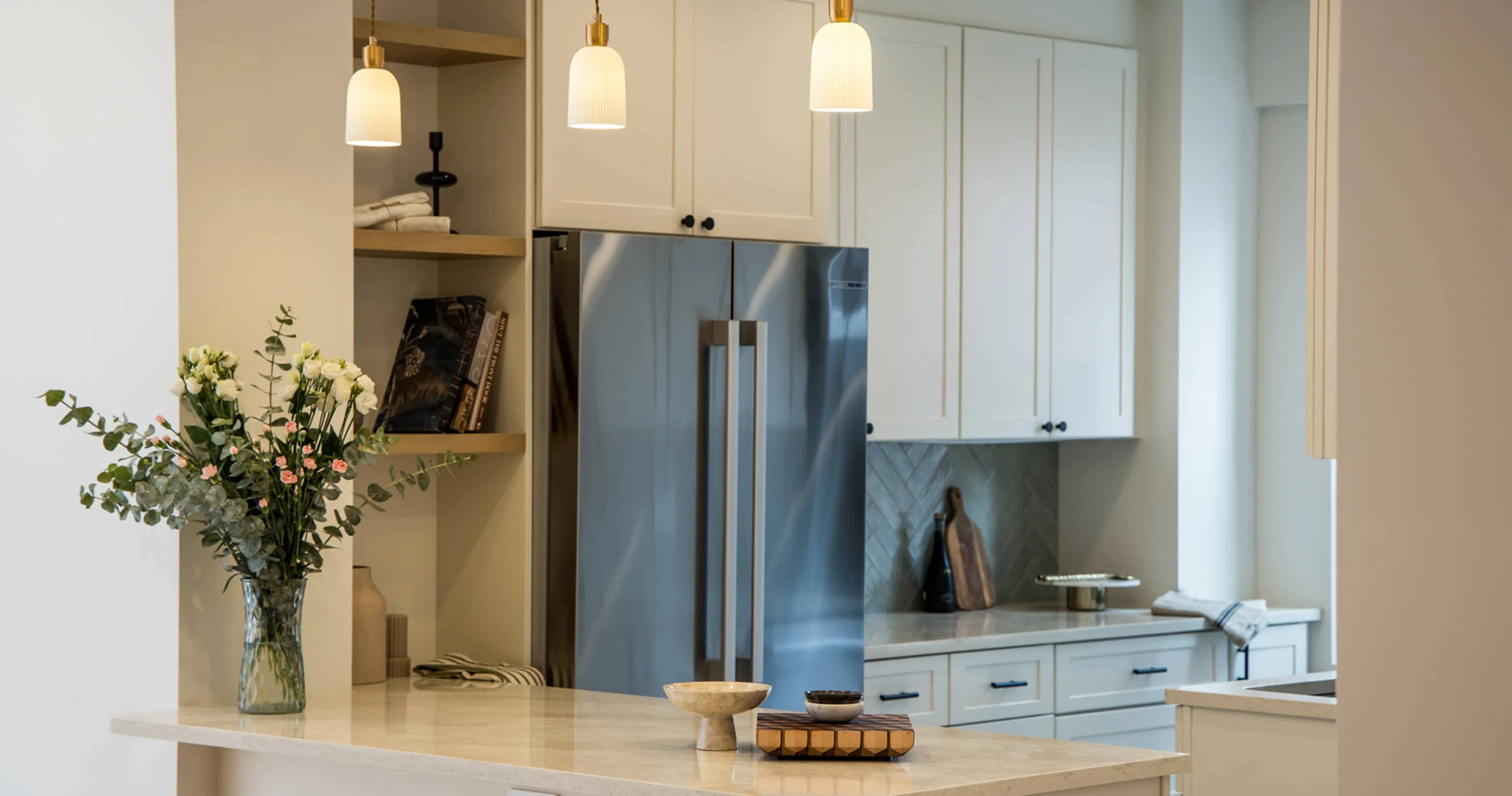 Close-up of kitchen bar area with open shelves, white shaker cabinets, brass pendant lights, stainless-steel refrigerator, and vase of flowers, part of a full apartment renovation at 80 East End Avenue in Yorkville, NYC