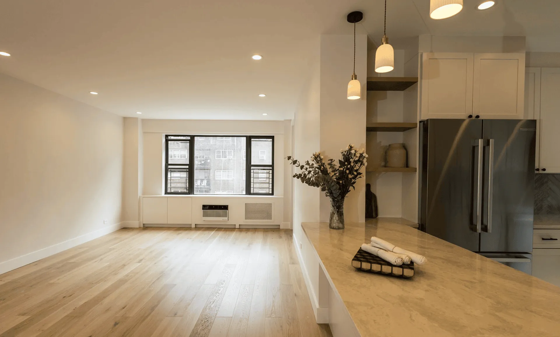 Open living and kitchen area after renovation featuring white waterfall island, pendant lights, wide‑plank oak floor, and large window, 80 East End Avenue Yorkville NYC