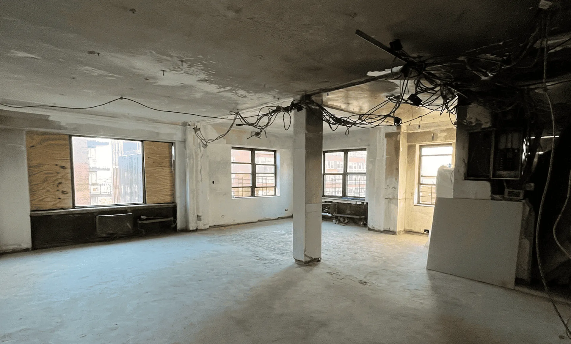 Another view of gutted apartment with exposed ceiling wiring, concrete floor, support column, and boarded windows before renovation, 80 East End Avenue Yorkville NYC
