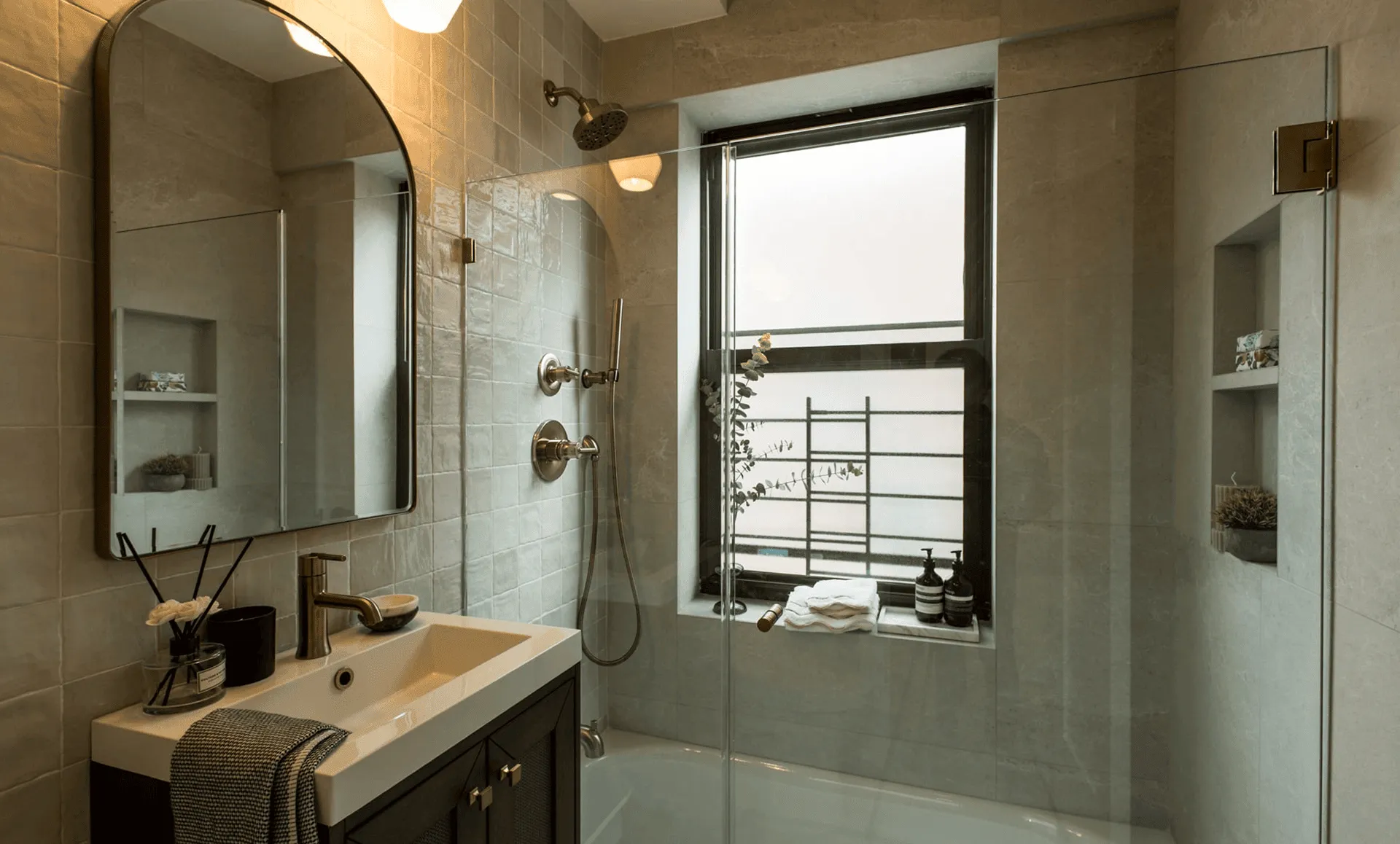 Modern bathroom with arched mirror, gray tile walls, built‑in niches, brushed‑nickel fixtures, and glass shower enclosure over bathtub, part of a full apartment renovation at 80 East End Avenue in Yorkville, NYC