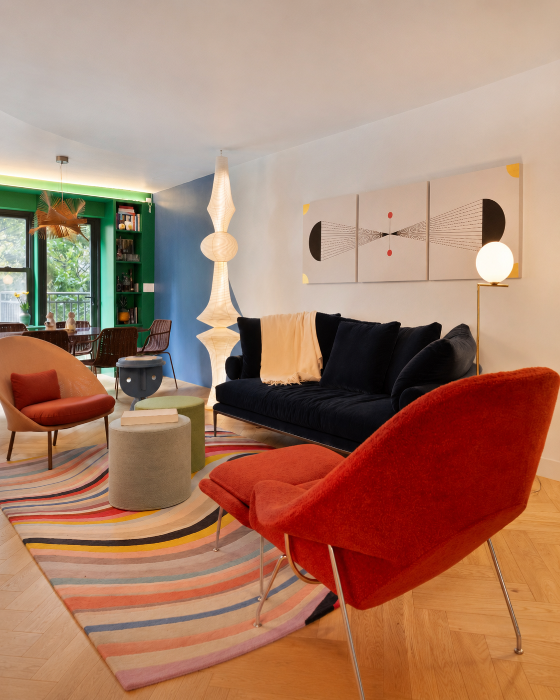 Renovated living room with a navy velvet sofa, red lounge chair, striped curved rug, sculptural white floor lamp, and a green dining nook beyond.