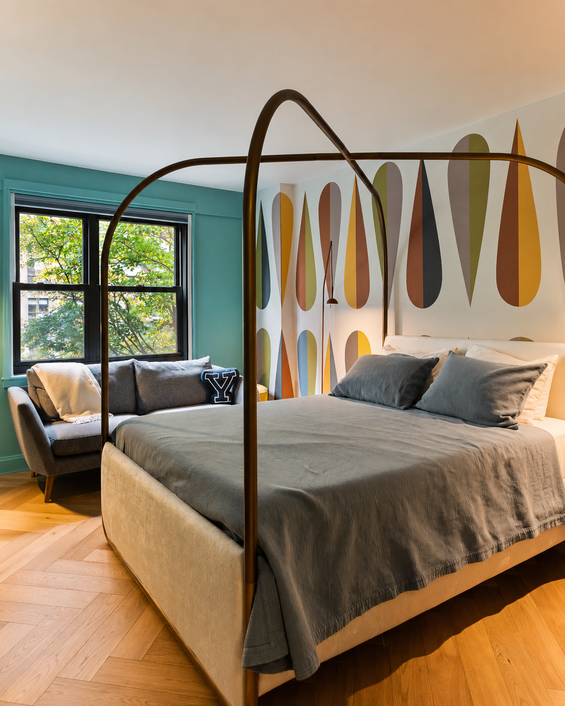 Renovated bedroom with a brass canopy bed, gray bedding, teal walls, a mid-century patterned accent wall, and herringbone wood floors.