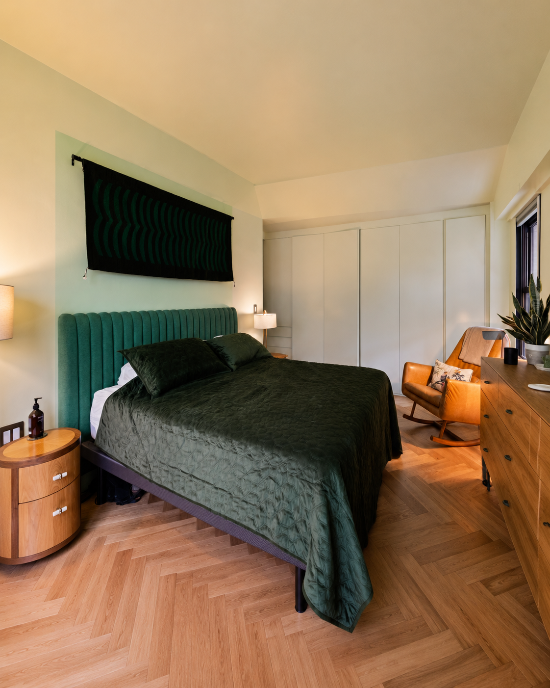 Renovated bedroom with a green upholstered bed, herringbone wood floor, tan leather rocking chair, wood dresser and nightstand, and built-in closets.