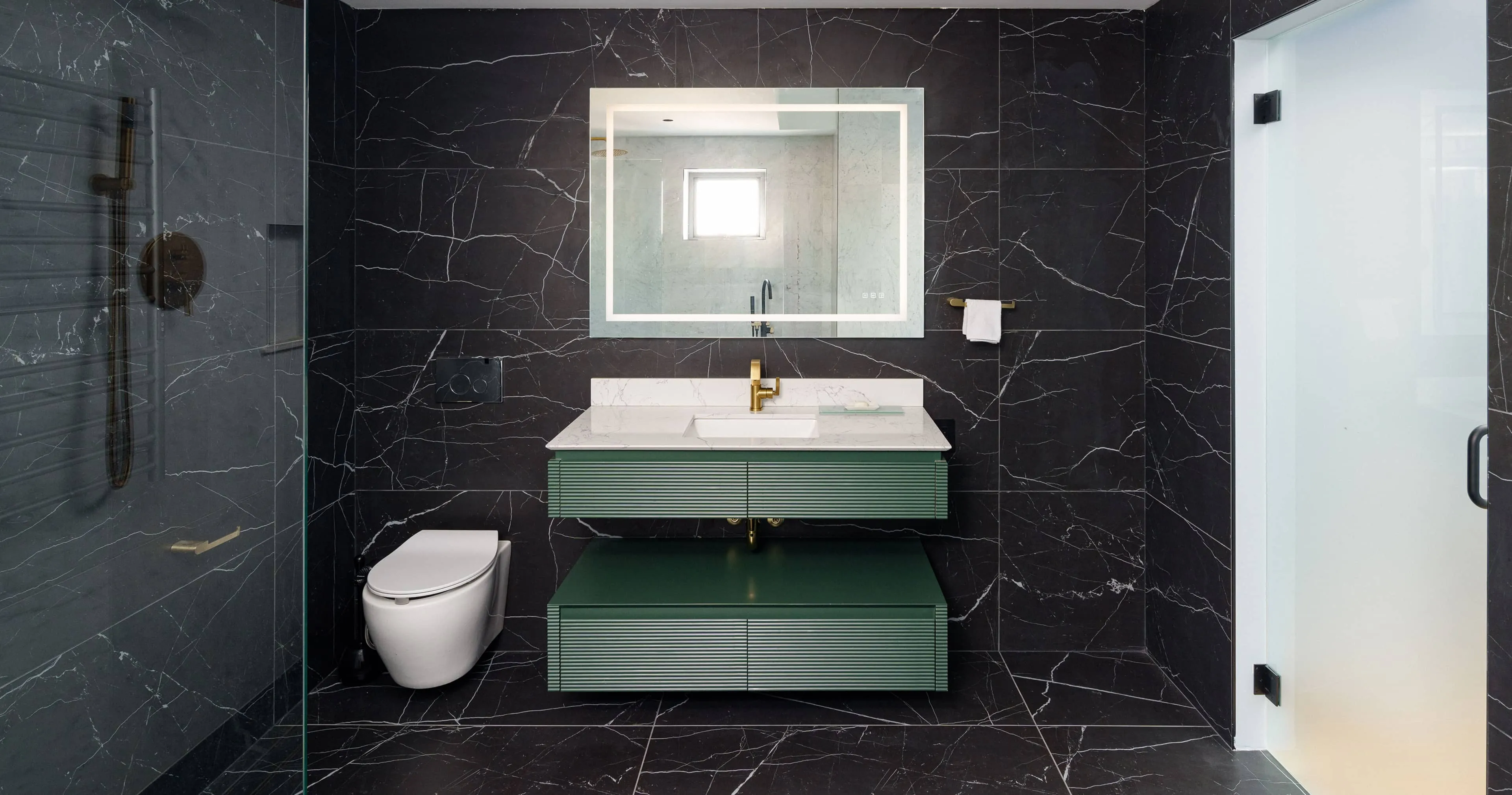 Full bathroom view with illuminated mirror, brass faucet, fluted green vanity with white marble countertop, wall‑hung toilet, black marble tile walls, and frosted glass door, part of an apartment renovation at 352 W 110th St Manhattan Valley NYC