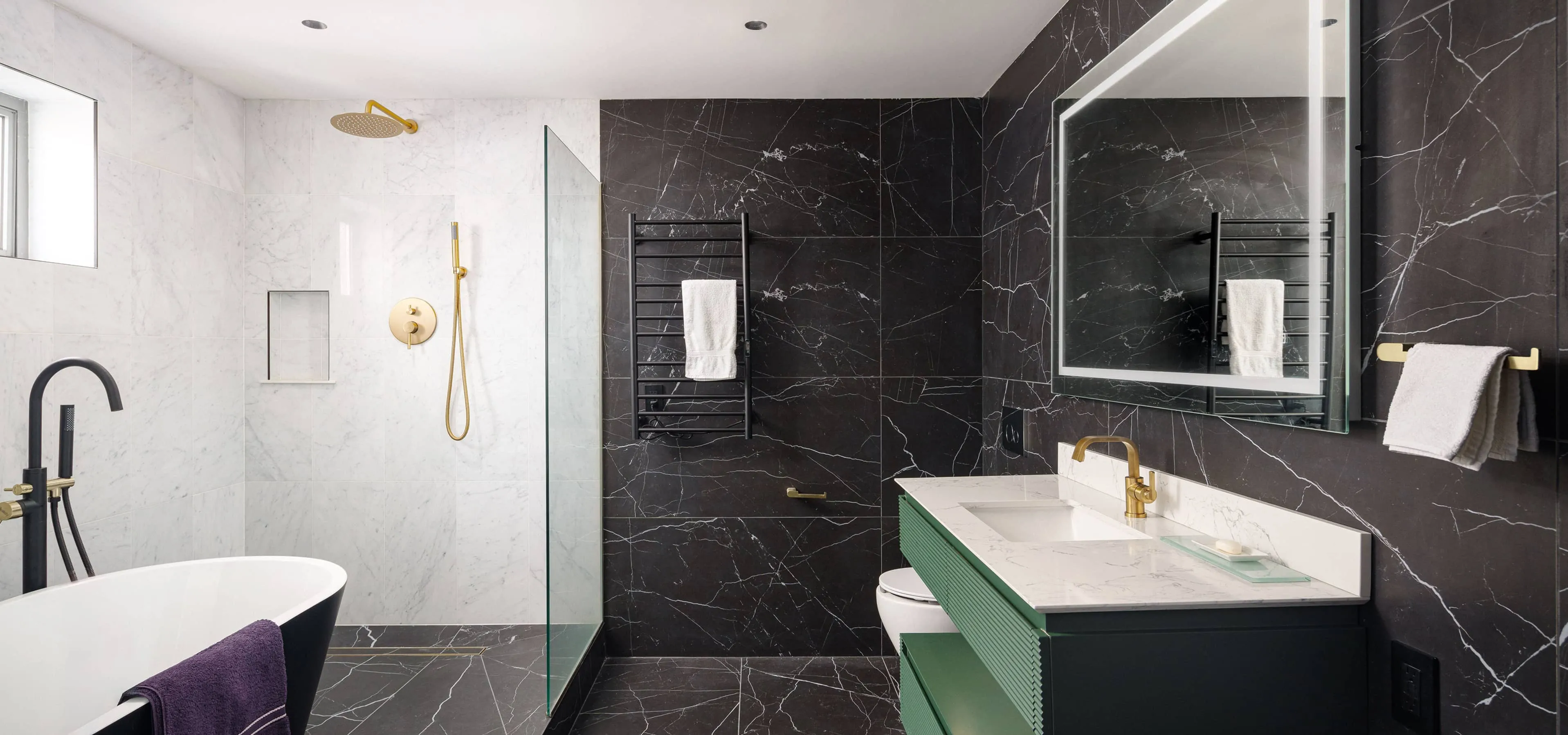 Modern bathroom featuring freestanding white tub with matte‑black faucet, glass‑enclosed shower with brushed‑gold fixtures, heated black towel rack, and green vanity with white marble countertop and brass faucet, part of an apartment renovation at 352 W 110th St in Manhattan Valley NYC