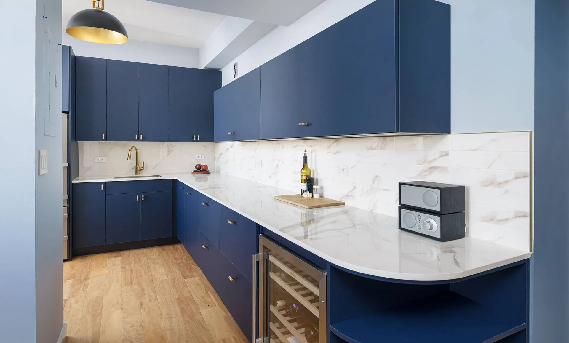 Kitchen with deep-blue flat-panel cabinets, white Dekton countertop and backsplash, oak flooring, and brass hardware, part of an apartment renovation at 352 W 110th St Manhattan Valley NYC