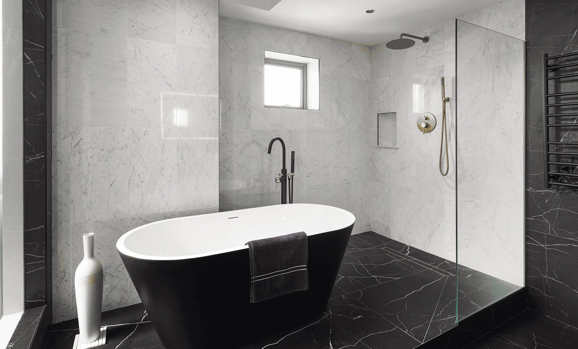 Spa-style primary bathroom with freestanding white tub, black marble floor and walls, brass shower and tub fixtures, glass partition, and black towel warmer, 352 W 110th St Manhattan Valley NYC